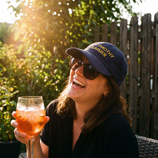 A woman outdoors in a sunny backyard setting, wearing a navy blue baseball cap with yellow embroidered text that reads 'JIMOTHY SUMMER' and holding a glass of Aperol Spritz.