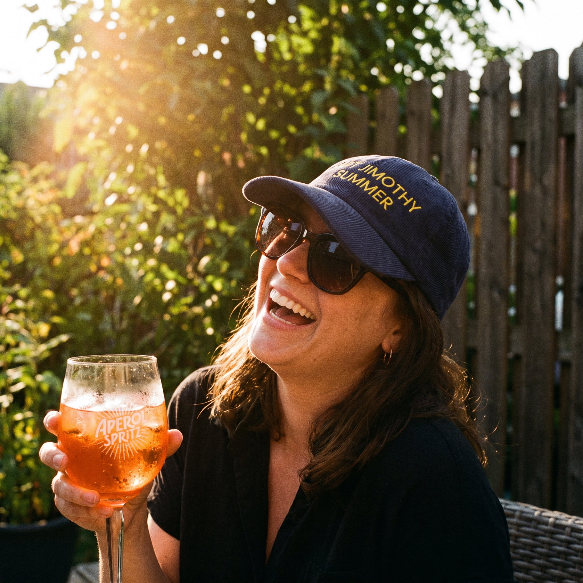 A woman outdoors in a sunny backyard setting, wearing a navy blue baseball cap with yellow embroidered text that reads 'JIMOTHY SUMMER' and holding a glass of Aperol Spritz.